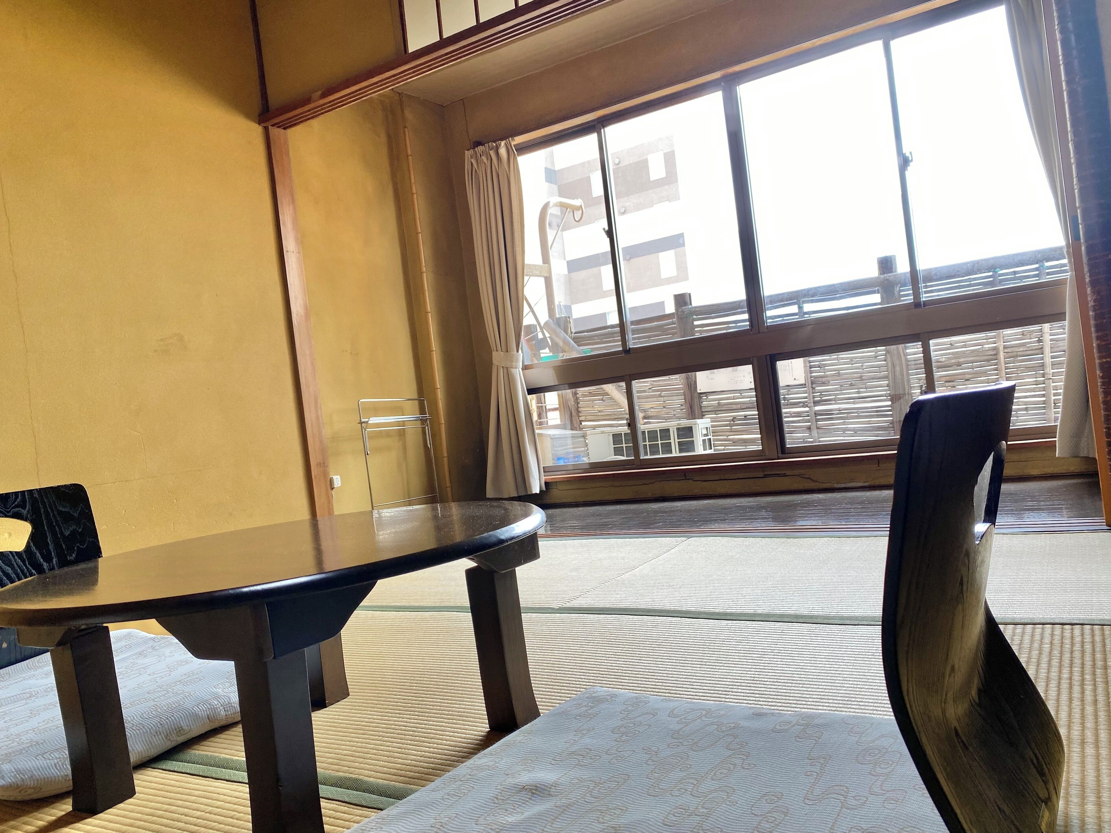A room with a tatami mat floor, a low table, a chair, and a large window with a view of buildings outside.
