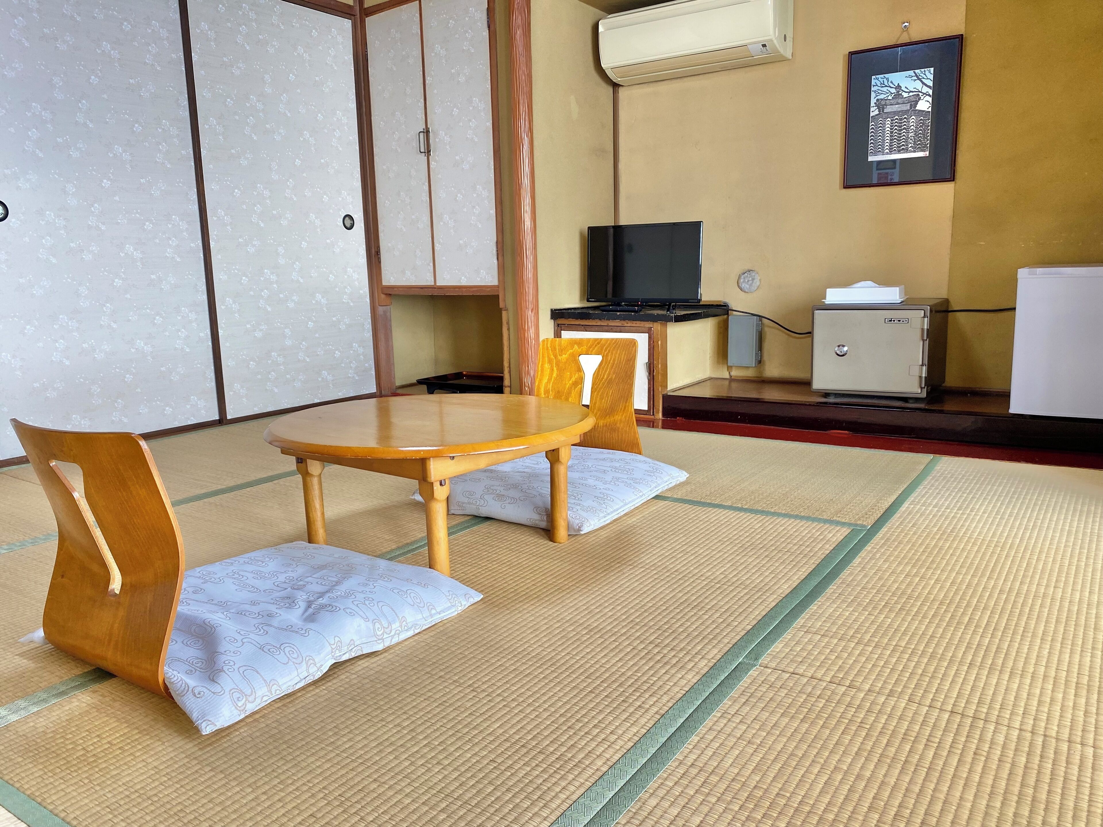 A room with tatami mat flooring, a wooden table with two chairs, a television, a cabinet, and a framed picture on the wall.