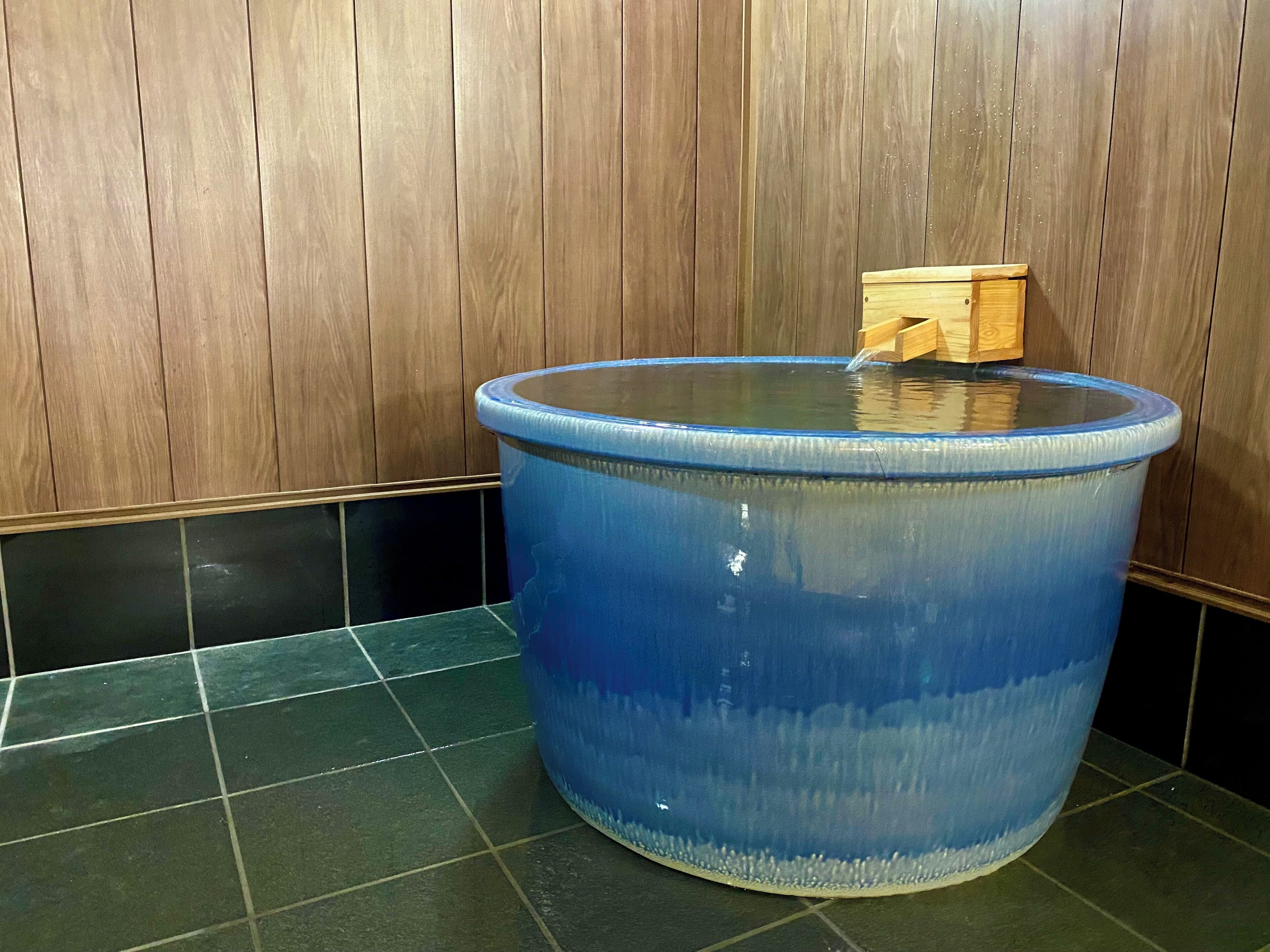 Un élément décoratif aquatique bleu, surmonté d’une structure en bois, installé devant un mur en bois et sur un sol carrelé.