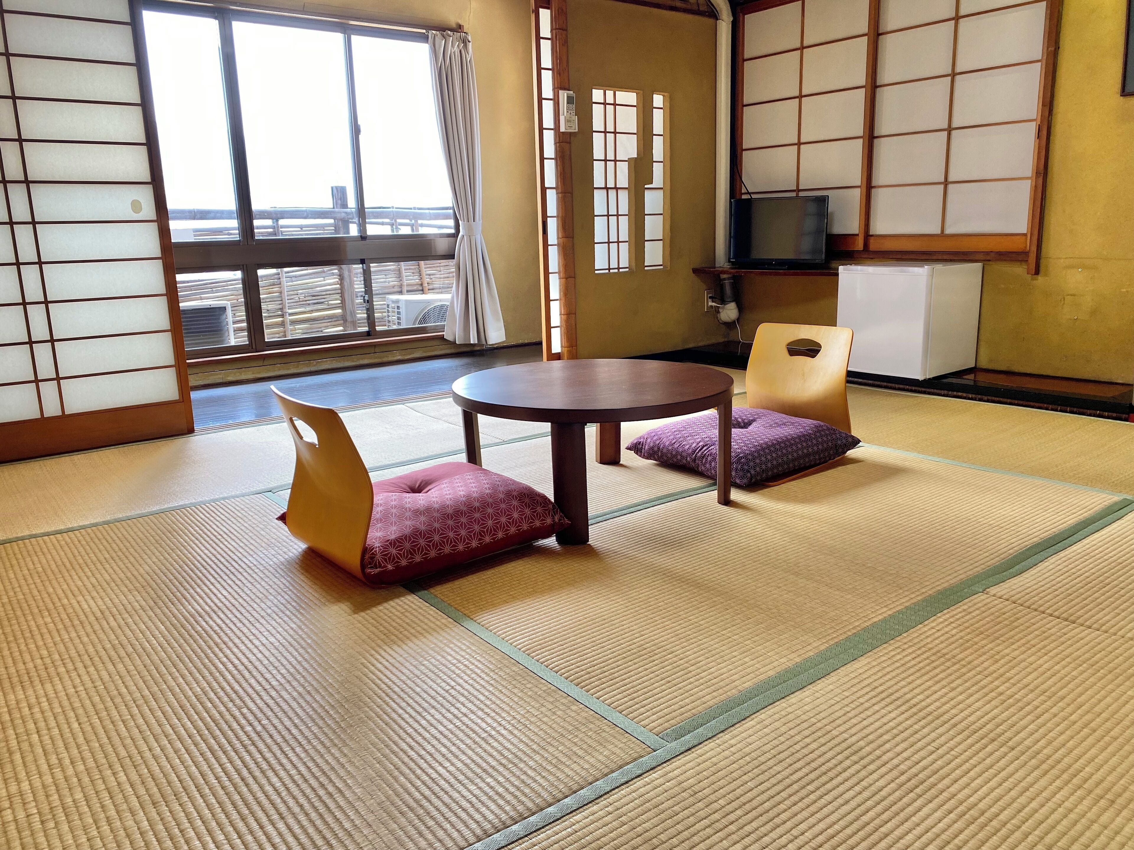 A traditional Japanese room with tatami flooring, a low table, two cushioned chairs, a television, and a view of the street outside.