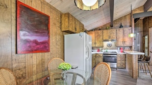 A wooden interior with a glass dining table, wicker chairs, a white refrigerator, wooden cabinets, and a large abstract painting. - Lake Arrowhead Cabin w/ Mid-century Charm! (Lake Arrowhead)