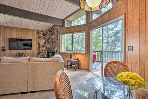 A cozy living room with a sofa, a TV, a stone wall, and large windows. - Lake Arrowhead Cabin w/ Mid-century Charm! (Lake Arrowhead)