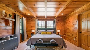 Ein Schlafzimmer in einer Blockhütte mit einem Bett, Nachttischlampen, einer Bank mit Handtüchern, einem Schminktisch und einem Fenster mit Vorhängen.