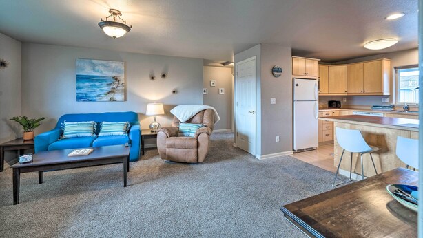 A living room with a blue sofa, a recliner, and a coffee table. There is a kitchen area with wooden cabinets and a refrigerator. - Apartment w/ Gas Fireplace ~ 1/2 Mi to Beach! (Anacortes)