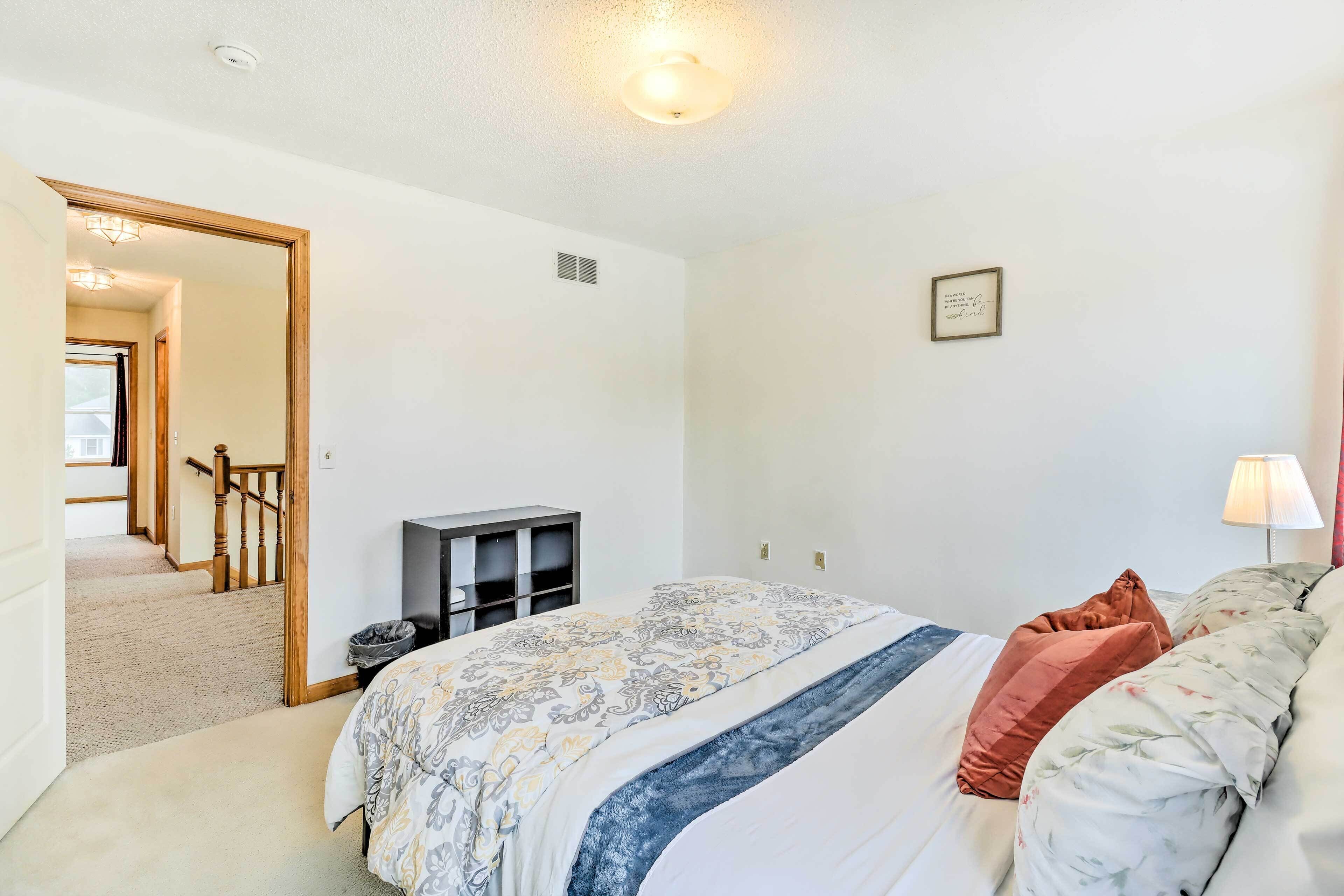 Un dormitorio con cama, mesita de noche, lámpara, una cita enmarcada en la pared y un pasillo que conduce a otra habitación.