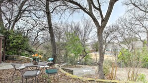 A patio area with a round table and chairs, surrounded by trees and a stone wall. - Studio w/ Pool - 13 Mi to Downtown Dallas! (Dallas)