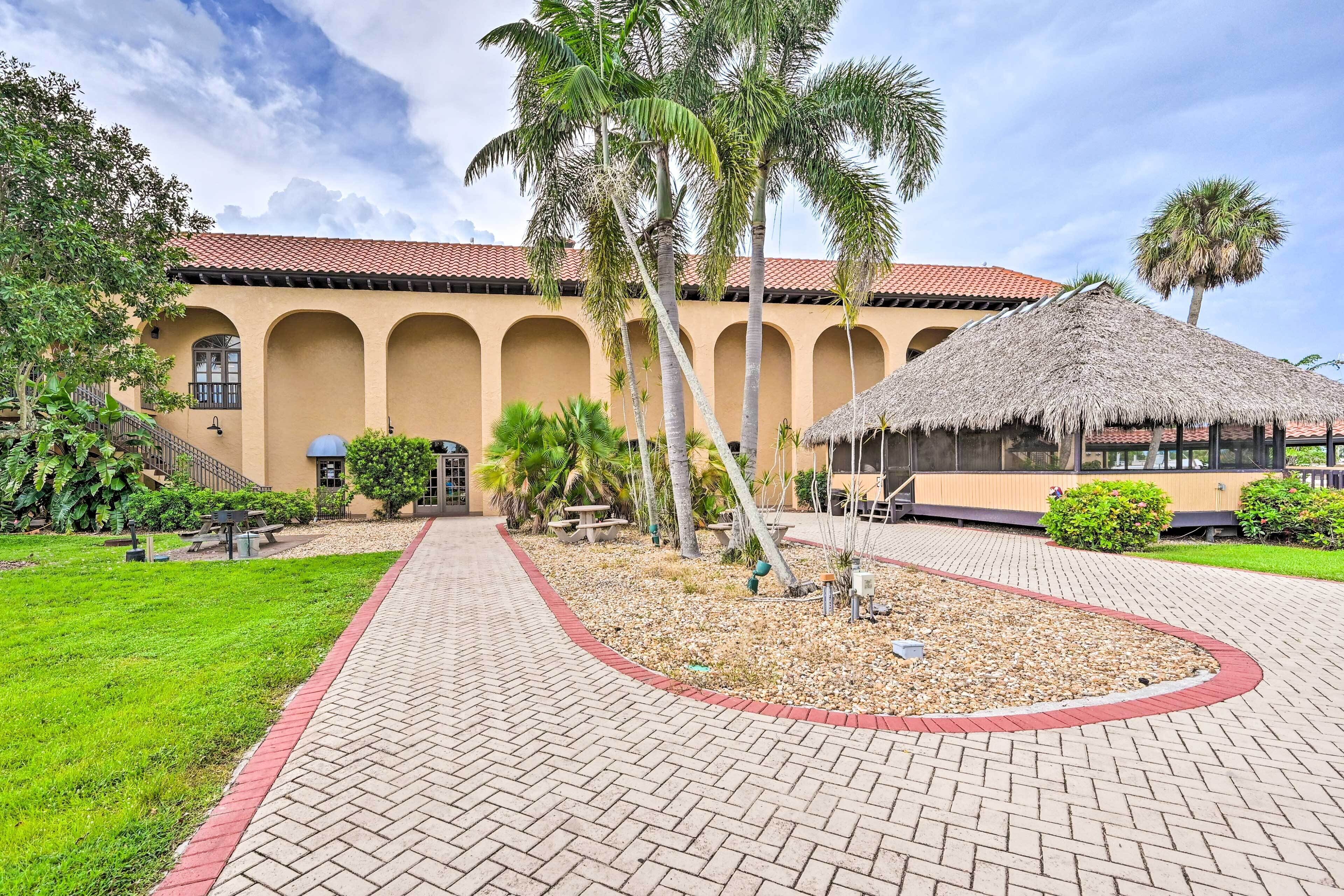 A courtyard with a paved walkway, palm trees, and a building with arched windows and a tiled roof.