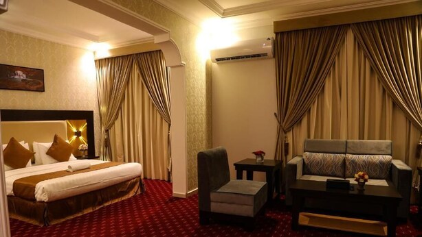 A hotel room with a large bed, a sofa, a small table, and a chair. There is a painting on the wall and an air conditioning unit on the wall. - Golden Rest Serviced Apartments (Al Khobar)