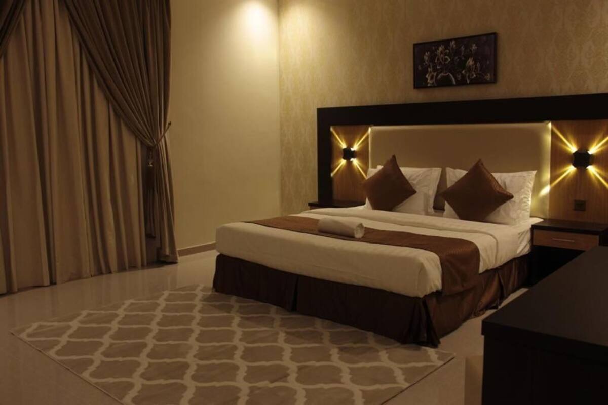 A hotel room with a large bed, a headboard with light fixtures, a nightstand, and a wall-mounted artwork.