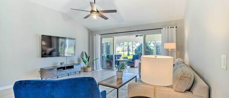 Una sala moderna con un sillón azul, un sofá, un televisor, un ventilador de techo y vistas al exterior.