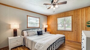 A bedroom with a wooden paneled wall, a bed with a white bedspread, two bedside lamps, and a ceiling fan. - Spacious Deck: Waterfront Home in Gun Barrel City! (Mabank)