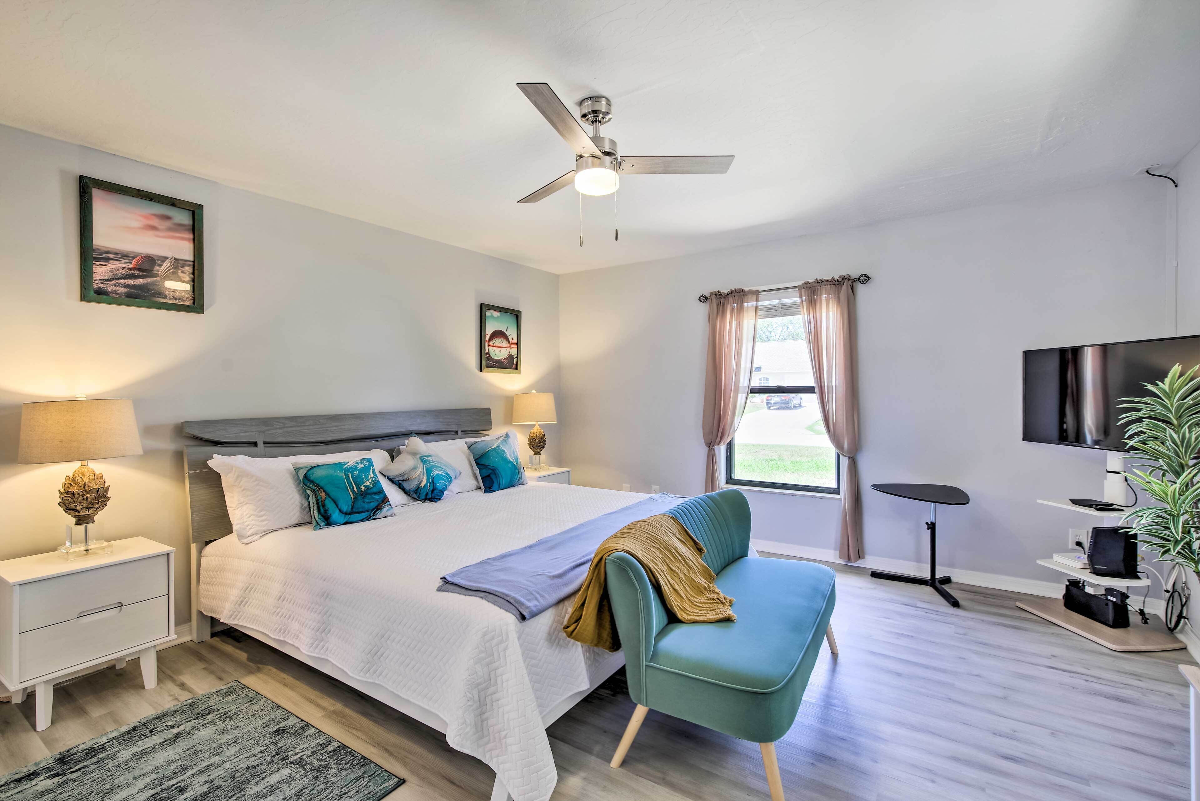 Une chambre à coucher comprenant un lit, des tables de chevet, un ventilateur de plafond, une fenêtre avec des rideaux, un téléviseur et une plante.