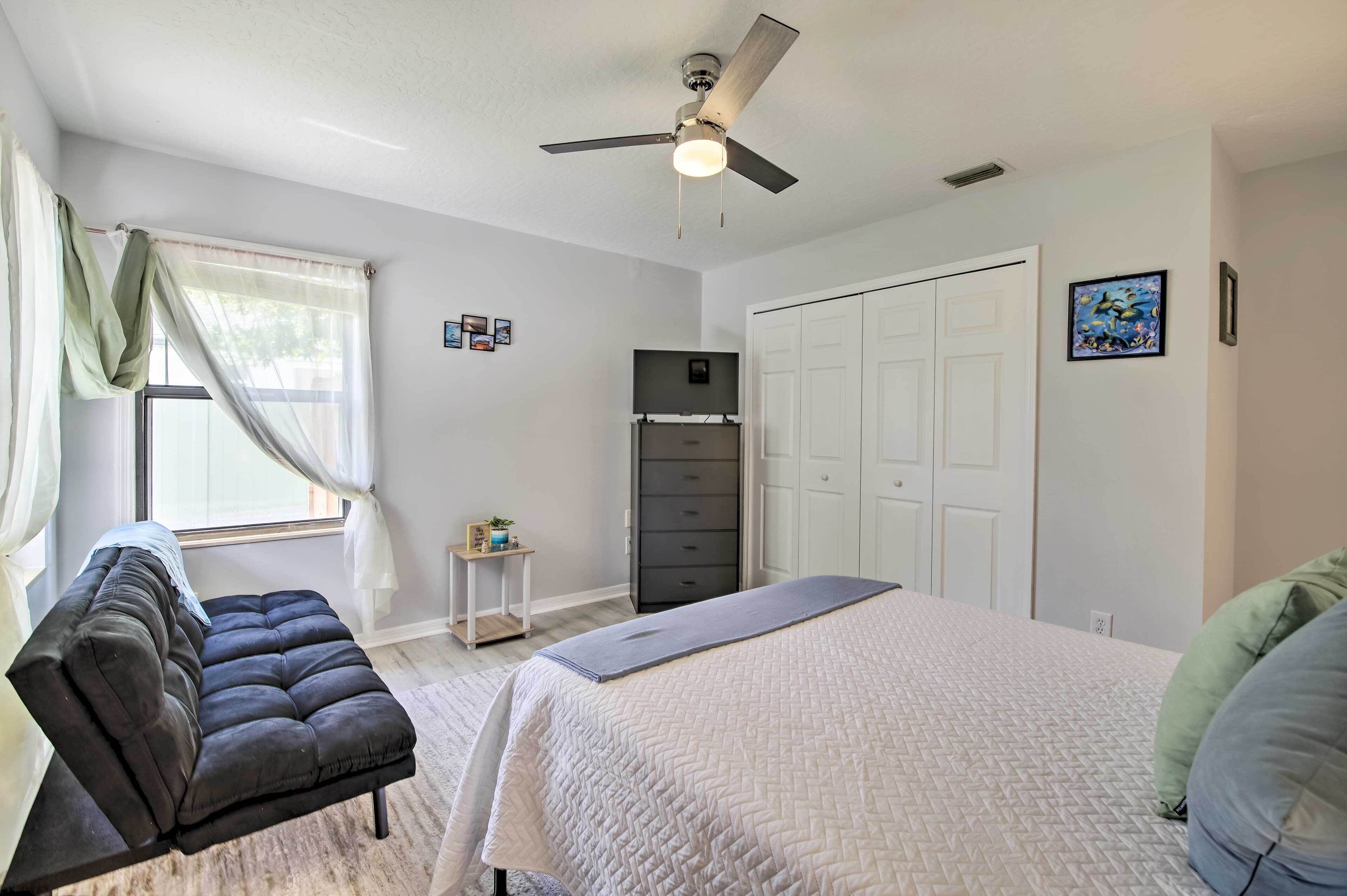 A bedroom with a bed, a dark sofa, a ceiling fan, a wardrobe, and a window with sheer curtains.