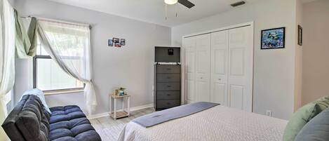 A bedroom with a bed, a dark sofa, a ceiling fan, a wardrobe, and a window with sheer curtains.