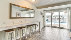 Une salle de bain moderne avec un comptoir en marbre, quatre tabourets, un grand miroir et une vue sur une piscine, visible à travers des portes vitrées.