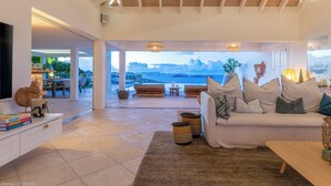Un espacio habitable amplio con un gran ventanal con vistas panorámicas, un sofá blanco con cojines, una mesa de centro de madera y un televisor de pantalla plana sobre un mueble blanco.