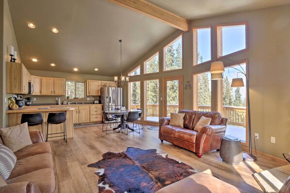 A spacious living area with a large wooden dining table, leather sofa, and a cowhide rug. There are multiple windows providing natural light and a view of the outdoors. - 26-0086 Fairplay Cabin w/ Hot Tub! (Fairplay)