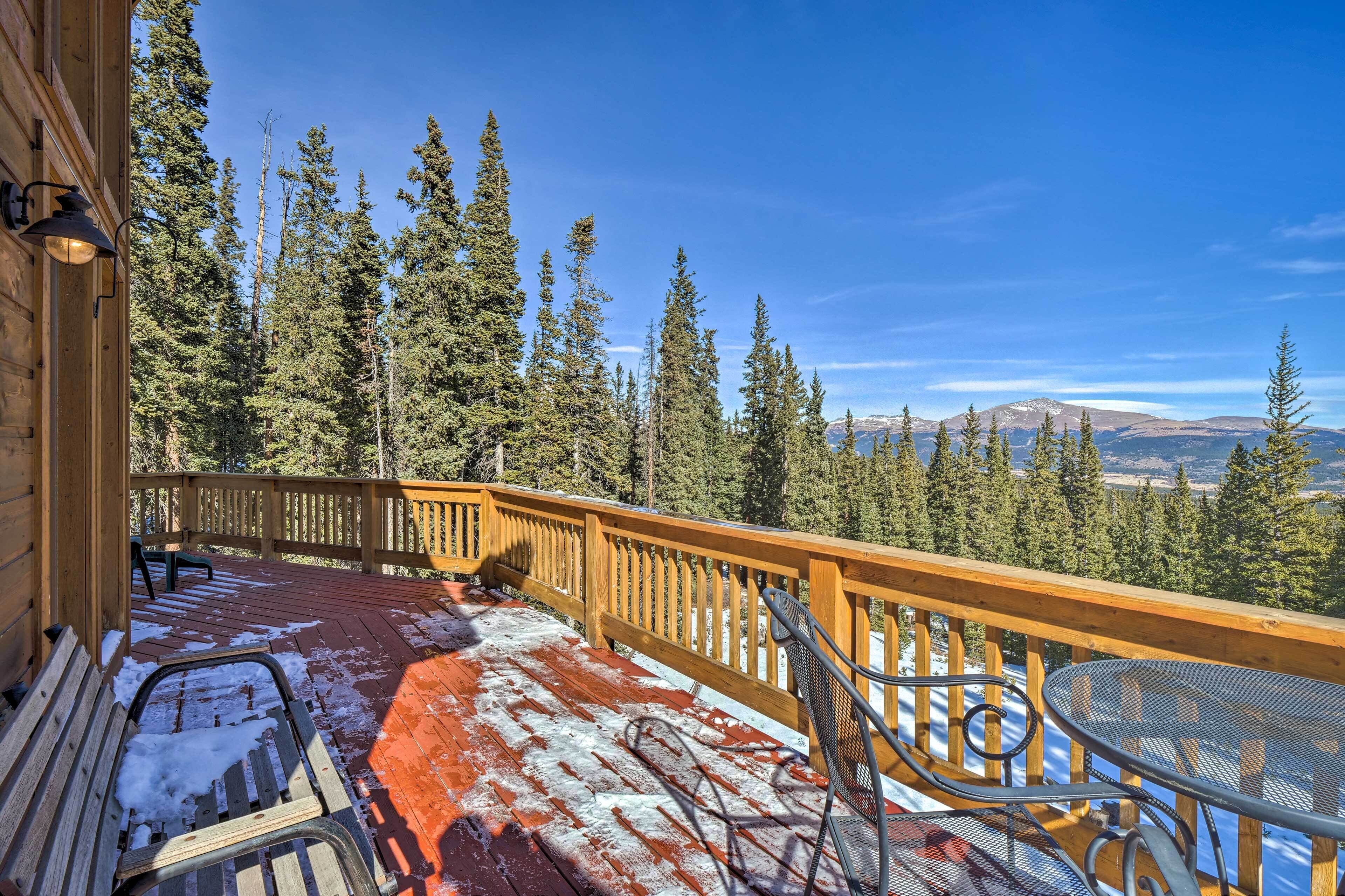 A wooden deck with a metal table and chairs, surrounded by a wooden railing, overlooking a forested mountain range.