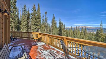 A wooden deck with a metal table and chairs, surrounded by a wooden railing, overlooking a forested mountain range.