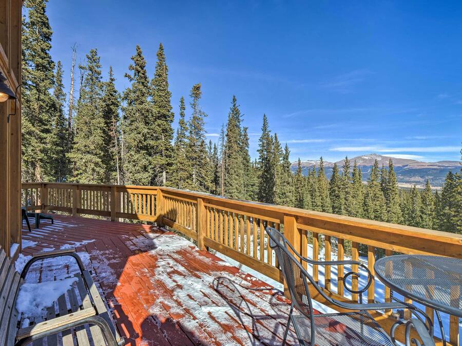 A wooden deck with a metal table and chairs, surrounded by a wooden railing, overlooking a forested mountain range.
