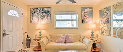 A living room with a beige sofa, glass coffee table, and two lamps. There is a ceiling fan, a door with a window, and artwork on the walls.