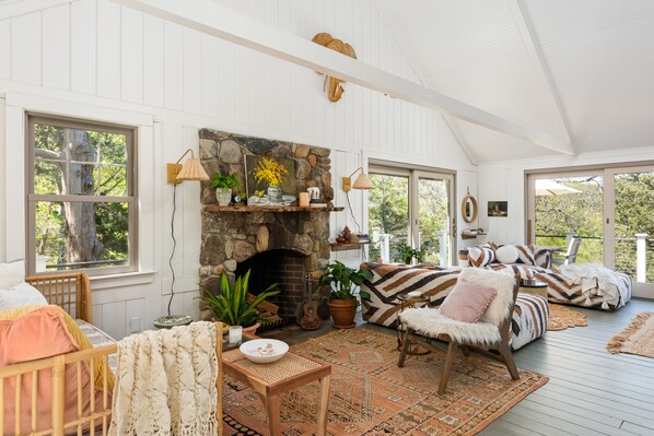 A cozy living room with a stone fireplace, wooden furniture, and large windows offering a view of the outdoors. - Peaceful Nauset Beach Sanctuary (Orleans)