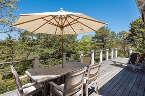 A wooden deck with a table and chairs, an umbrella, and a railing overlooking a forested area. - Peaceful Nauset Beach Sanctuary (Orleans)