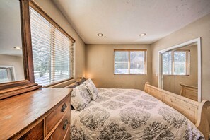 A bedroom with a wooden dresser, a large bed with patterned bedding, and two windows with blinds. - Family-friendly Sagle Vacation Rental w/ Deck (Sagle)