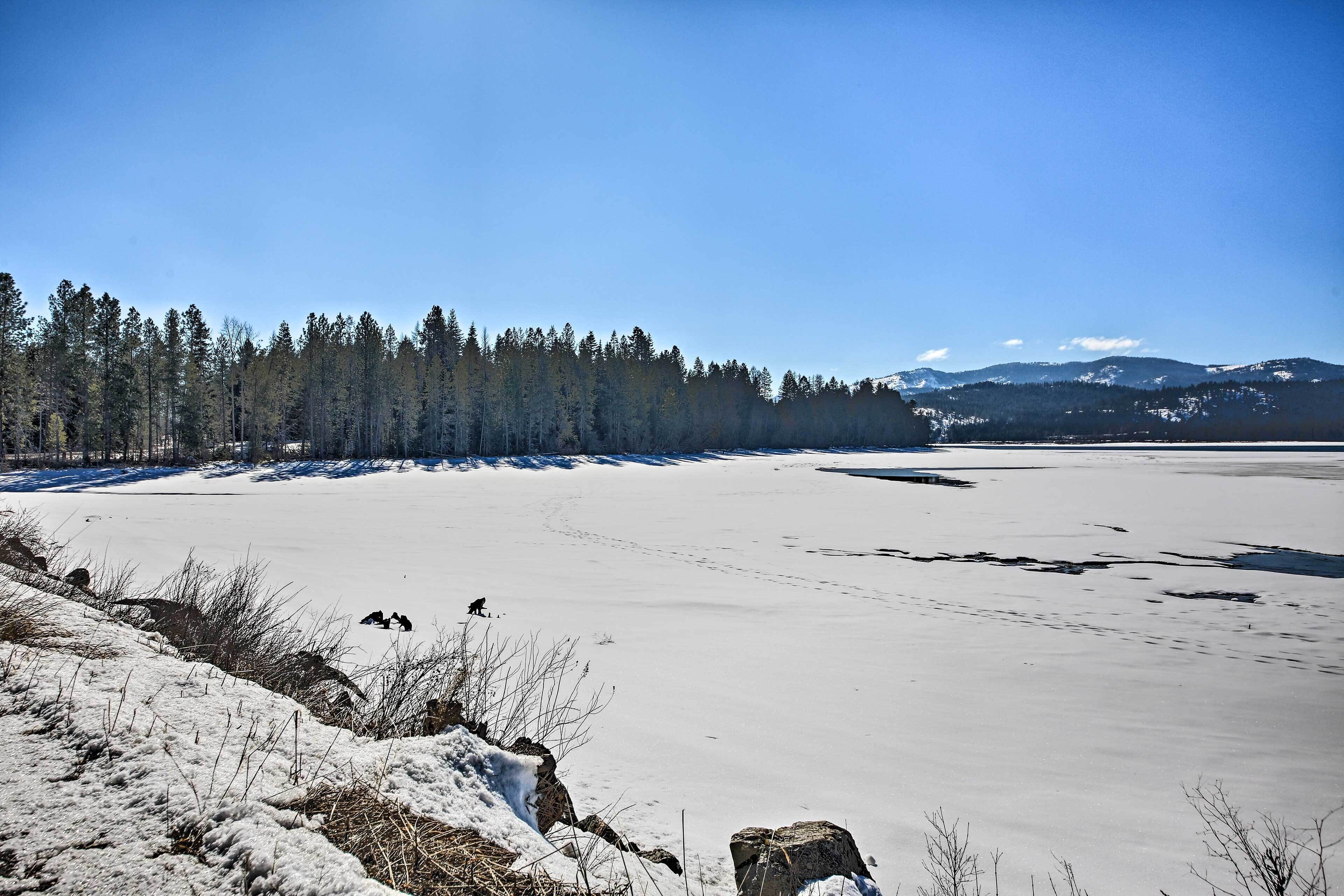 A snow-covered landscape with a frozen lake, trees, and mountains in the background.