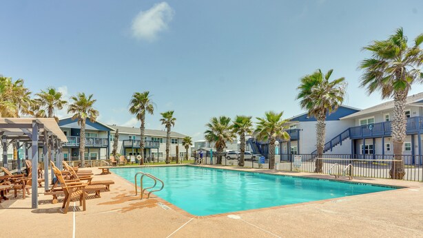 A swimming pool with lounge chairs, palm trees, and a building with balconies. - 'sea-king Sol' Ocean-view Condo in Port Aransas! (Port Aransas)