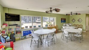 A dining area with a round table and chairs, a television mounted on the wall, and a view of the street through large windows. - 'sea-king Sol' Ocean-view Condo in Port Aransas! (Port Aransas)