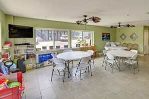 A dining area with a round table and chairs, a television mounted on the wall, and a view of the street through large windows. - 'sea-king Sol' Ocean-view Condo in Port Aransas! (Port Aransas)