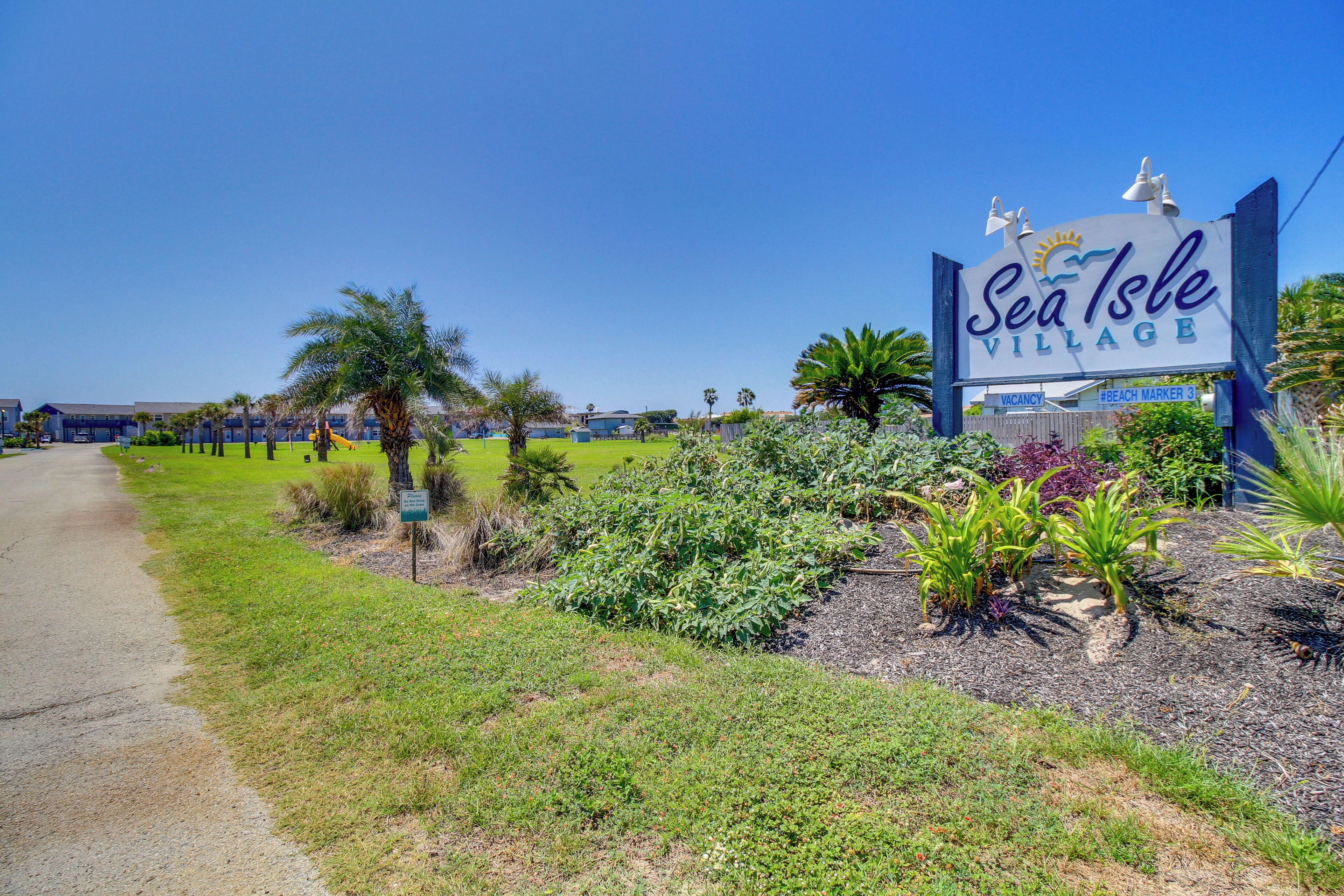 Señal de Sea Isle Village con cielo despejado y zona verde con plantas.