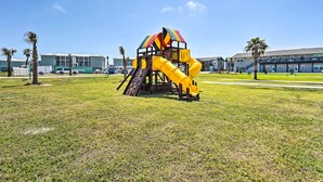 A playground with a slide and climbing structure, surrounded by grass and palm trees, with buildings in the background. - 'sea-king Sol' Ocean-view Condo in Port Aransas! (Port Aransas)