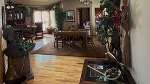 A spacious living area with a high ceiling, wooden flooring, and a dining area with a table and chairs. There is a large window providing natural light, and a ceiling fan is visible.