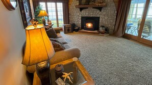 A cozy living room with a stone fireplace, a ceiling fan, and a view of the outdoors through large windows.
