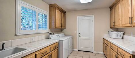 A kitchen with wooden cabinets, a built-in washer and dryer, a window with blinds, and a white door.