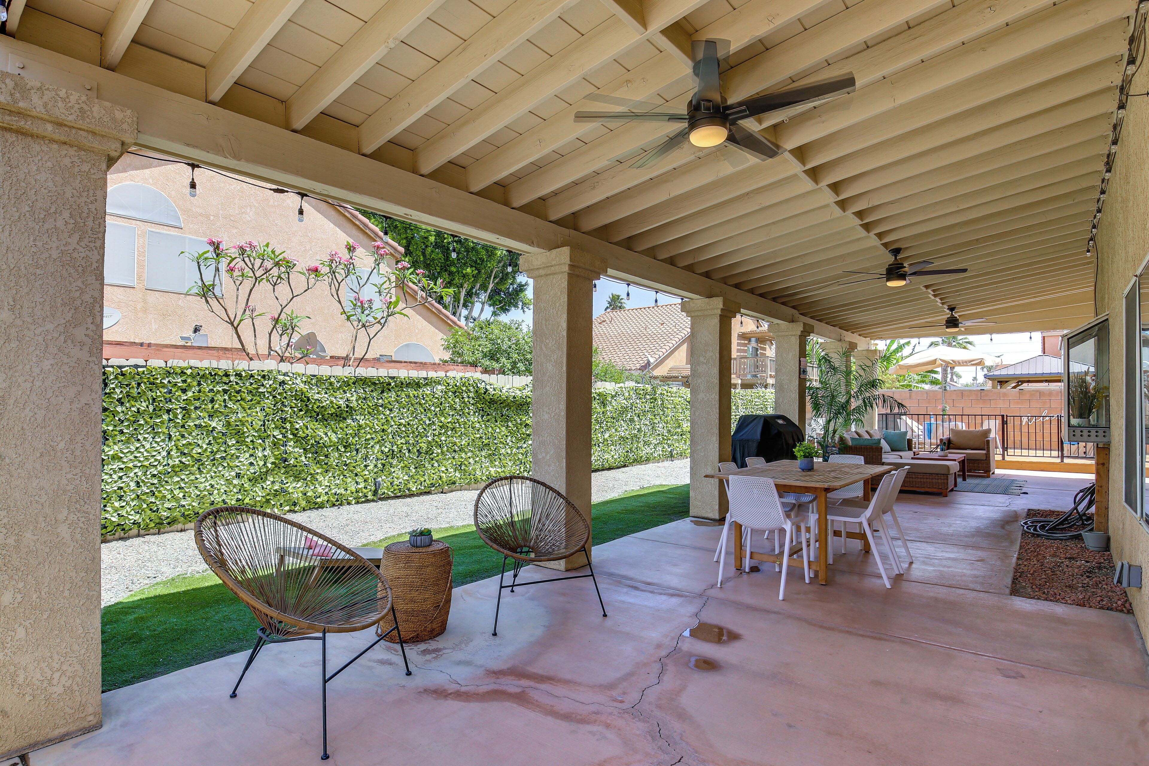 Une terrasse couverte avec un sol en béton, des ventilateurs de plafond et du mobilier d’extérieur comprenant des chaises et une table.