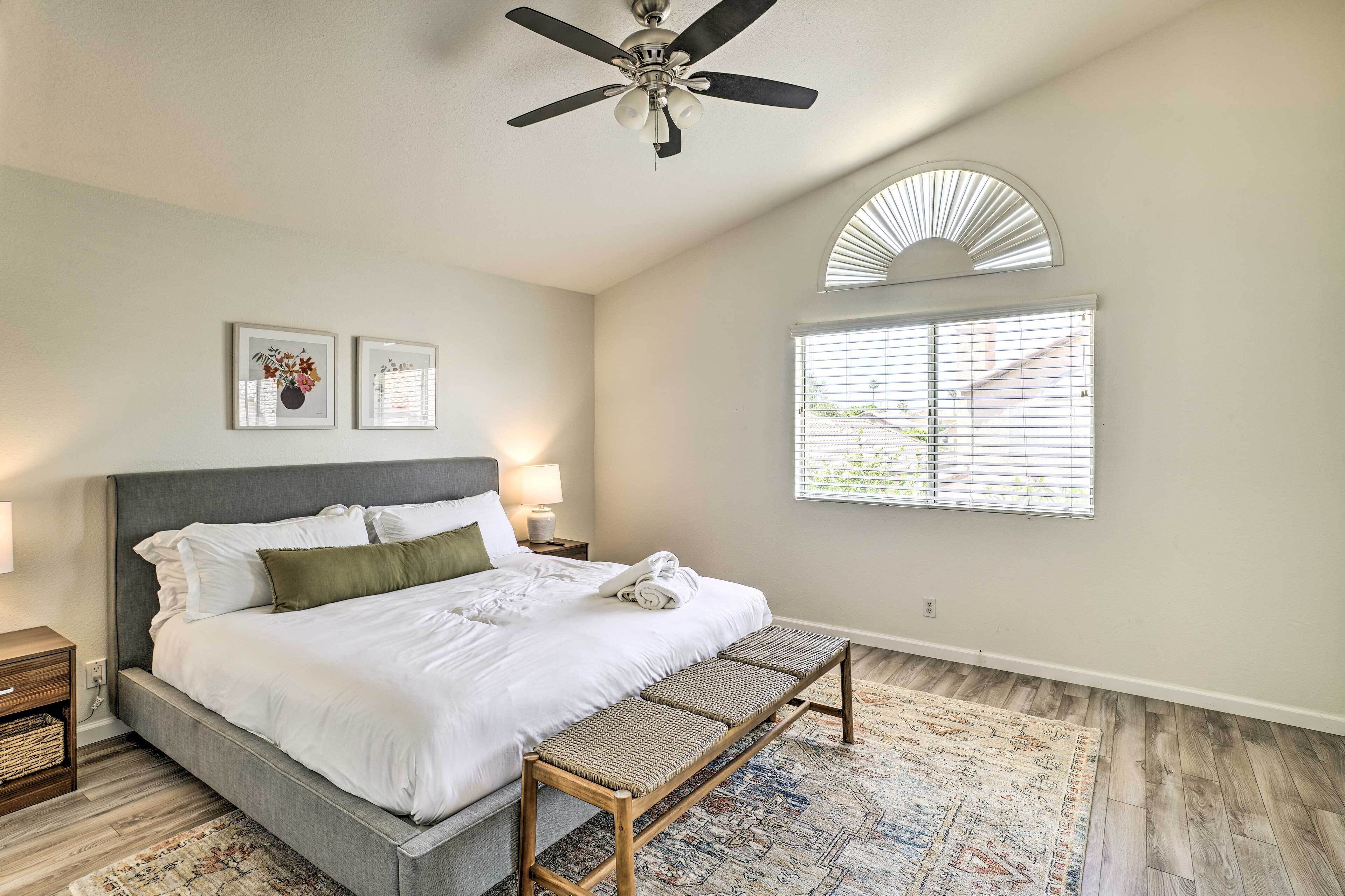 A bedroom with a bed, a bench, a ceiling fan, a window with blinds, and two framed pictures on the wall.