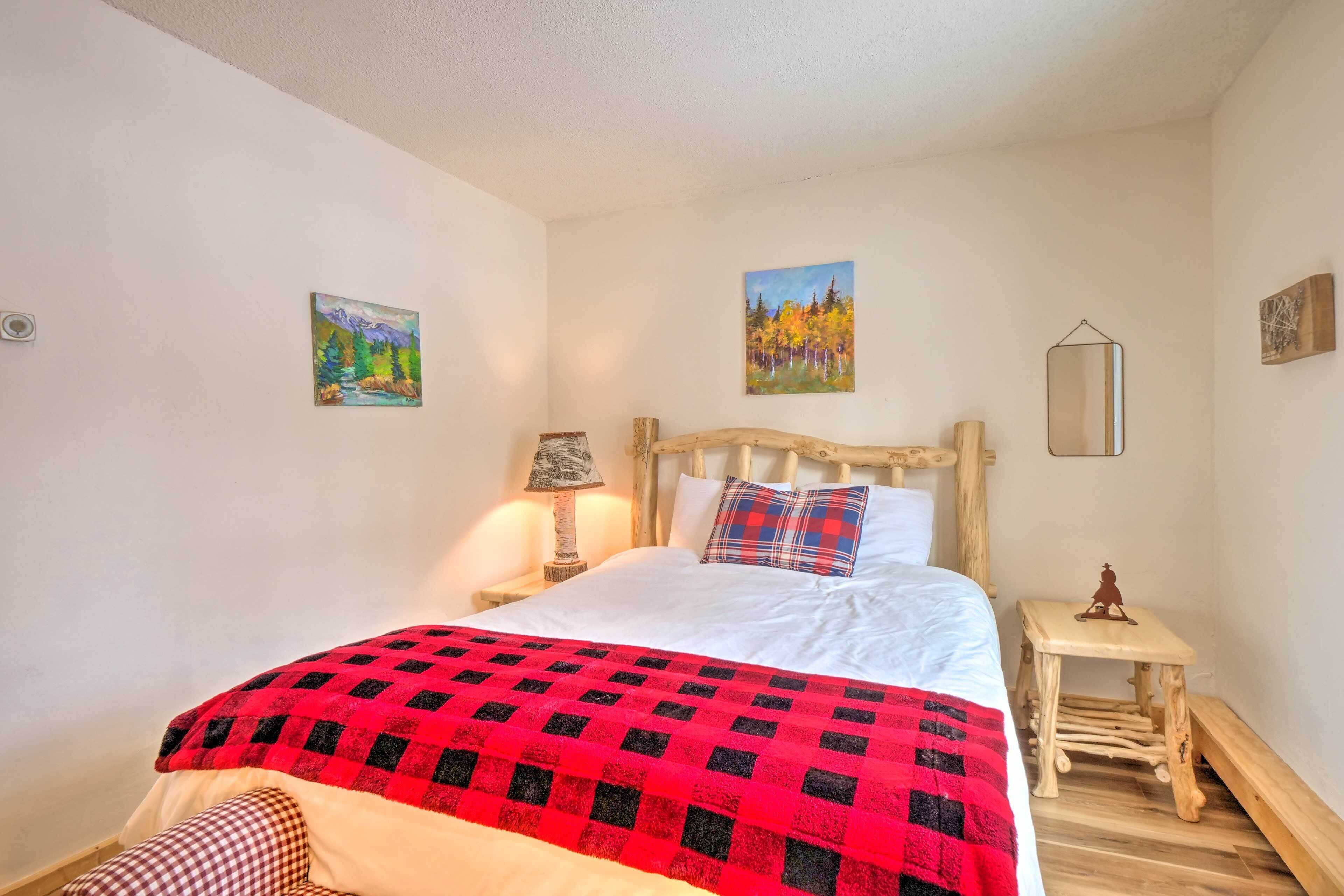 Une chambre avec un lit en bois, une couverture à carreaux rouges et noirs, deux tableaux accrochés au mur, une lampe de chevet et une petite table de nuit en bois.