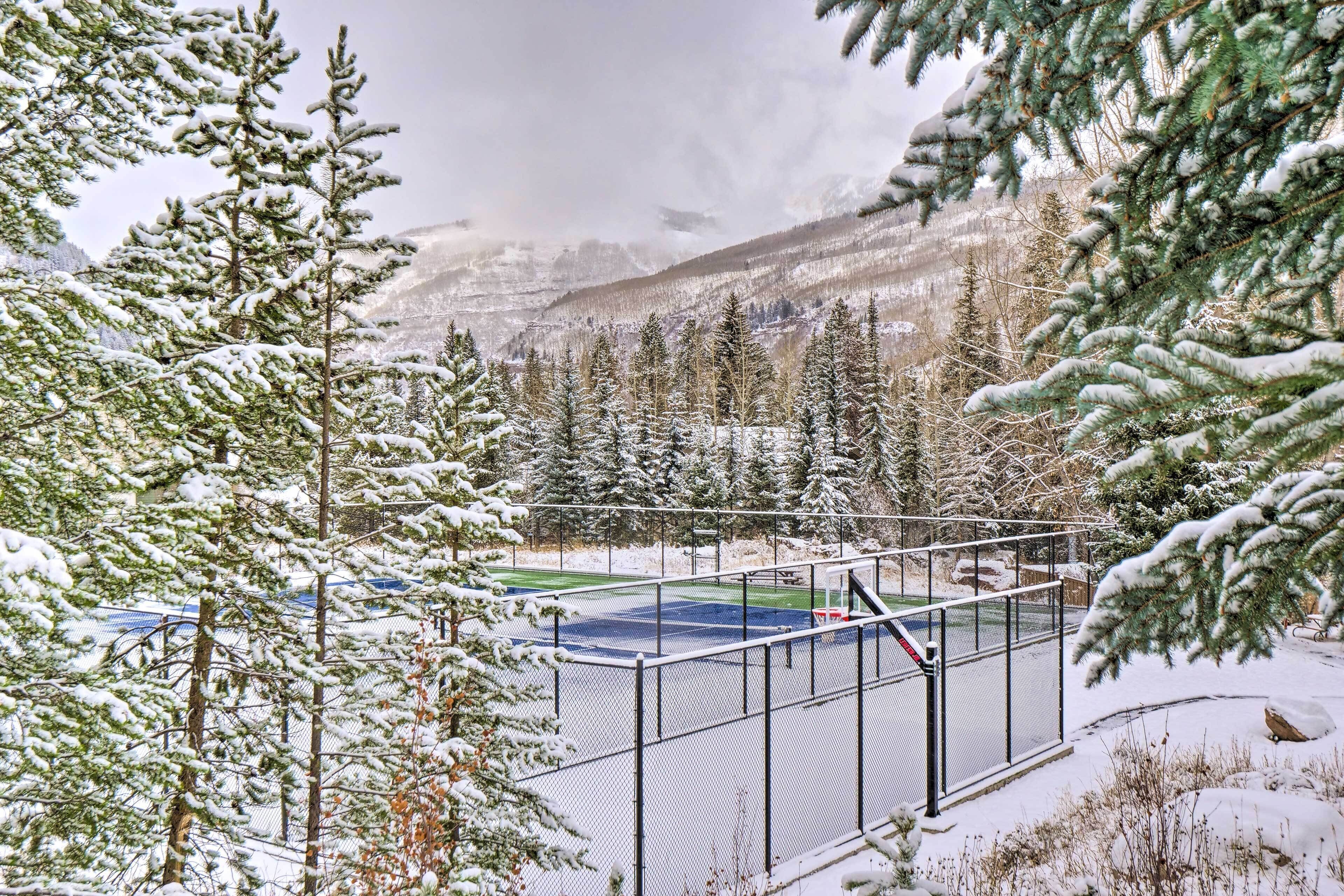 Un área exterior nevada con una cancha de tenis cercada, rodeada de árboles de hoja perenne y montañas al fondo.