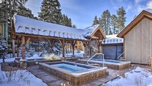 A wooden spa area with two hot tubs and a covered seating area, surrounded by snow and trees.