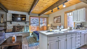 A spacious living area with a fireplace, a large TV mounted on a stone wall, and a kitchen with white cabinets.