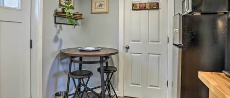 A compact kitchen with a refrigerator, a small dining table with stools, a door with a decorative panel, and a trash can.