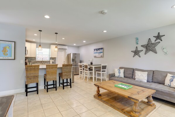 A modern kitchen with white cabinets, a dining area with wicker chairs, a wooden coffee table, and a gray sofa with decorative pillows.