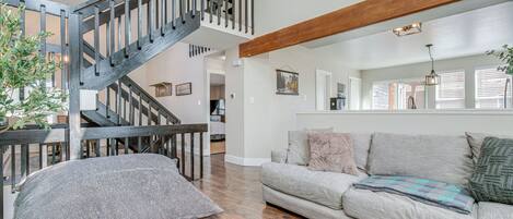 A spacious living room with a wooden staircase, a grey sofa, and a large window with blinds.