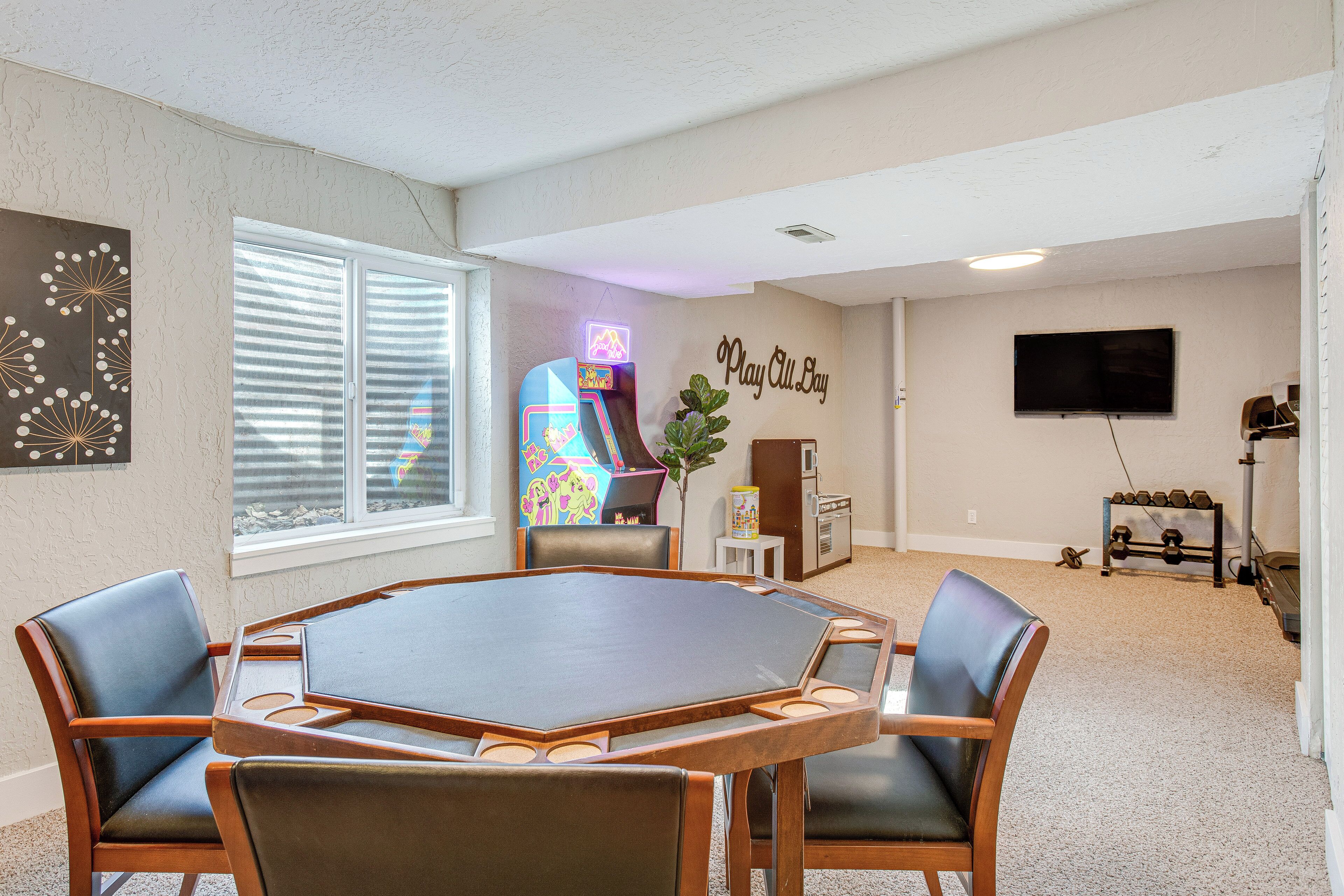 A room with a round table and four chairs, a neon arcade machine, a television, and a wall-mounted dumbbell rack.