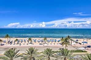 A beach with rows of blue umbrellas and palm trees, a road in the foreground, and the ocean in the background.