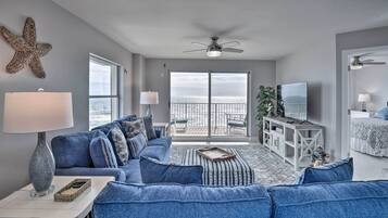 A modern living room with a blue sofa set, a white side table, a ceiling fan, and a view of the ocean through large sliding doors.