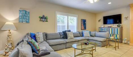 A living room with a sectional sofa, a glass coffee table, a ceiling fan, and a television on the wall.